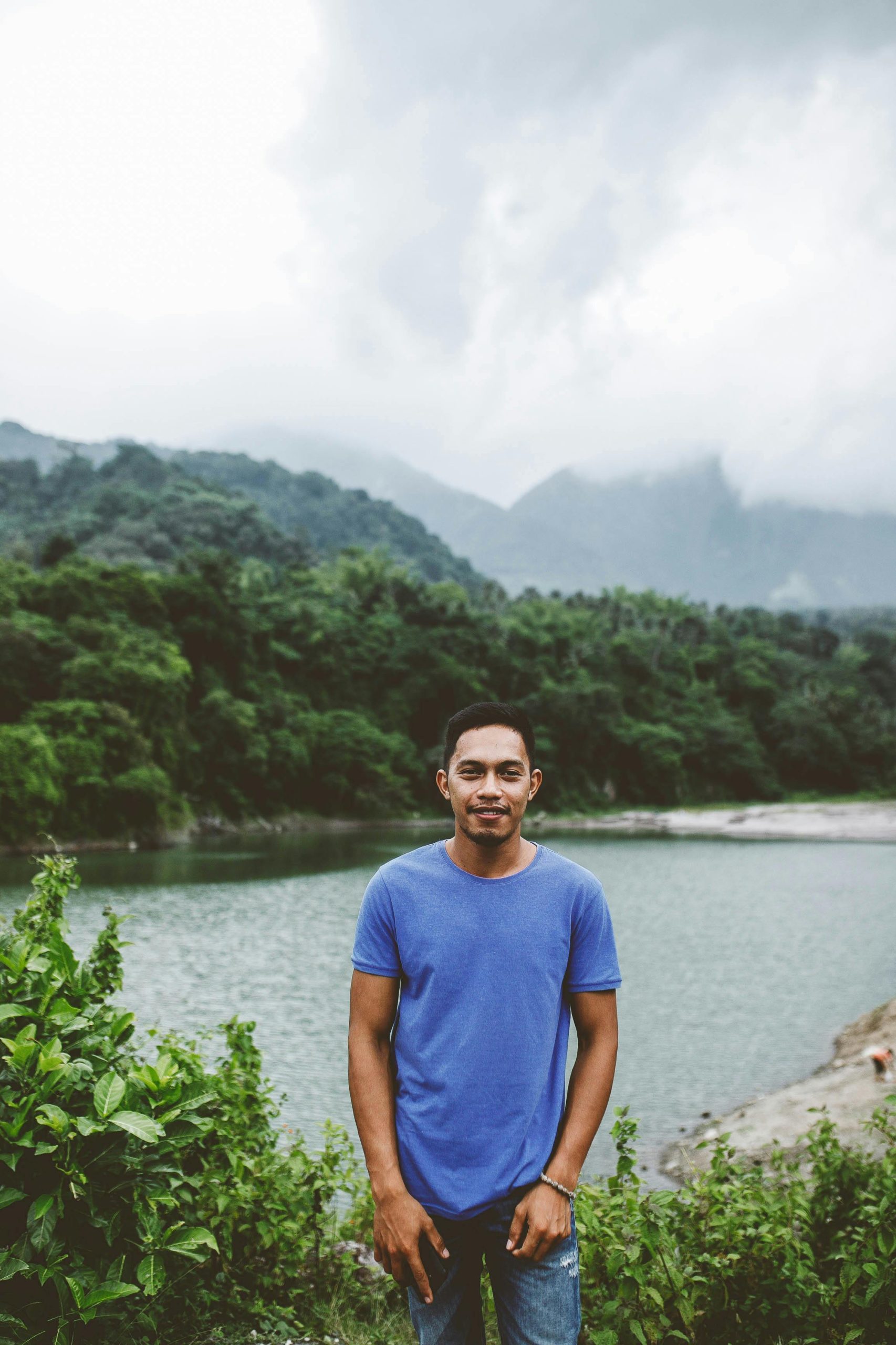 A man in a blue shirt stands smiling by a lake, surrounded by misty mountains and lush greenery.